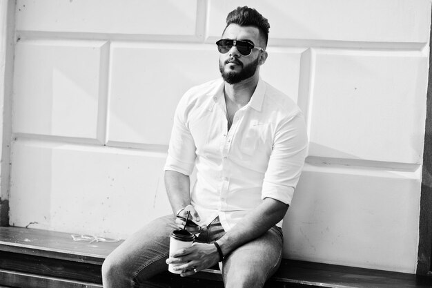 Stylish tall arabian man model in white shirt jeans and sunglasses posed at street of city and hold mobile phone at hand Beard arab guy with cup of coffee sitting and having rest