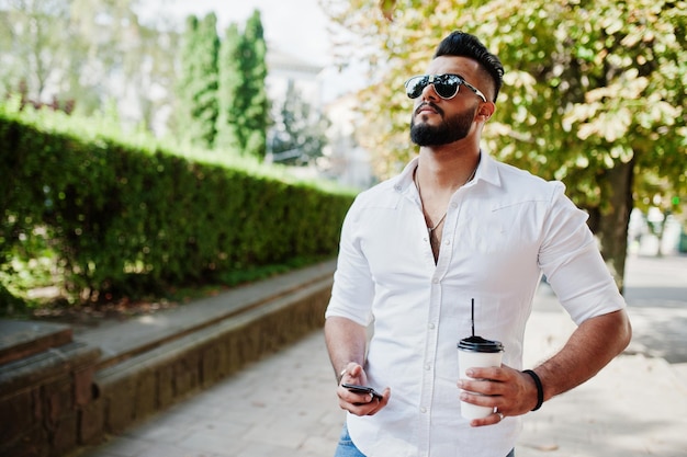 흰색 셔츠 청바지와 선글라스를 입은 세련된 키 큰 아라비아 남자 모델은 손에 커피 한 잔과 휴대 전화를 들고 도시 수염 매력적인 아랍 남자의 거리를 걷고