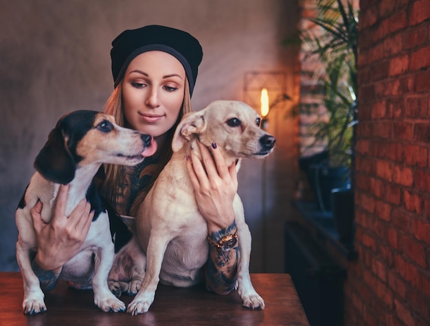 A stylish tattoed blonde female in t-shirt and jeans embraces two cute dogs.