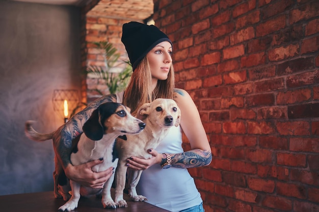 Стильная татуированная блондинка в футболке и джинсах обнимает двух милых собак.
