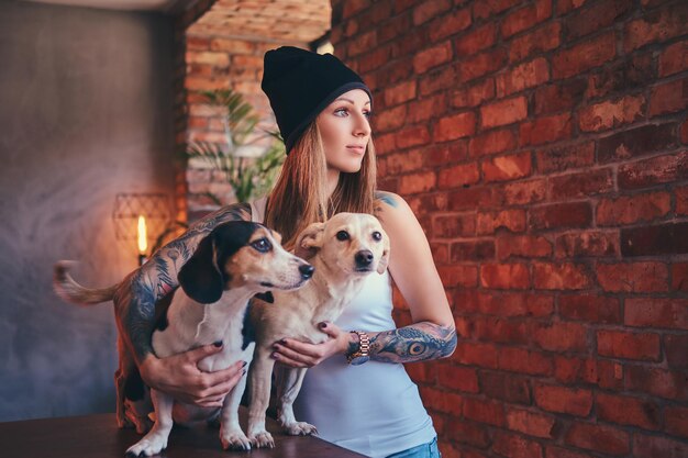 A stylish tattoed blonde female in t-shirt and jeans embraces two cute dogs.