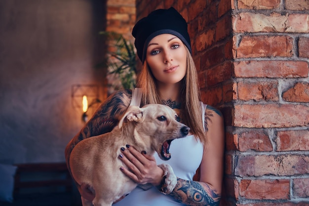 A stylish tattoed blonde female in t-shirt and jeans holds a cute dog.
