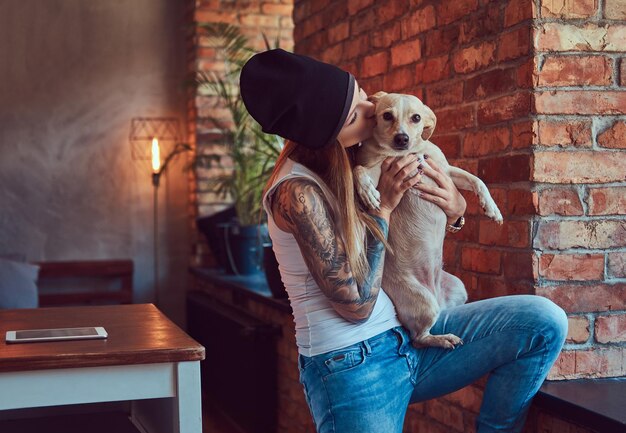 Tシャツとジーンズのスタイリッシュなtattoedブロンドの女性はかわいい犬を保持します。