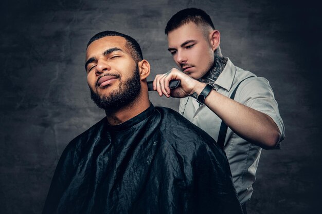 Stylish tattooed barber makes haircut to a Black bearded male.