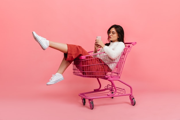 キュロットと白いセーターのスタイリッシュな十代の少女は、スーパーマーケットのトロリーに座っています。メガネのモデルがキスをし、ピンクで自分撮りをします。