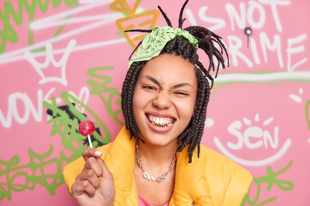 Stylish teenage girl clenches teeth has fun with friends poses against graffiti wall holds lollipop has trendy hairstyle wears metal chain around neck yellow vest