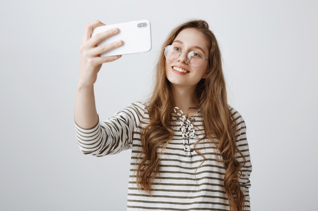Stylish teenage girl taking selfie on smartphone, smiling happy