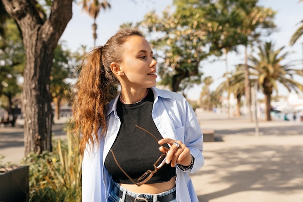 Stylish trendy girl with waving hair wearing black top and blue shirt is holding sunglasses and looking aside while walking on sunny street with exotic plants on background