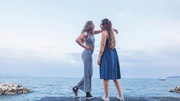 Stylish two young female friends standing against blue sea and sky