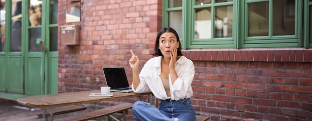 Free photo stylish urban girl asian woman with laptop sits in cafe points at upper right corner banner shows