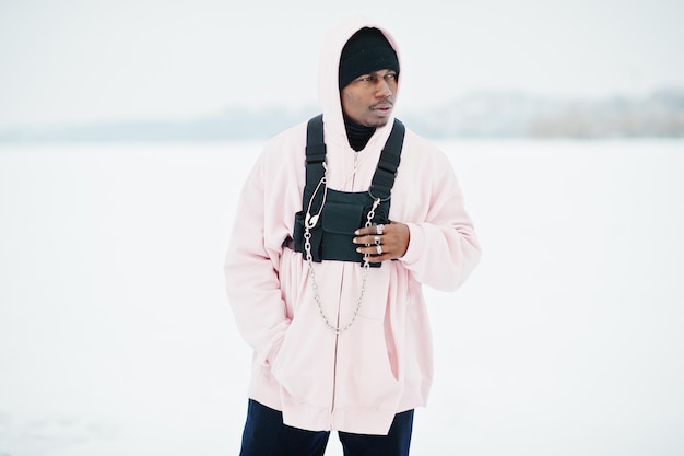 Elegante uomo afroamericano in stile urbano con felpa con cappuccio rosa posato sul lago ghiacciato in inverno