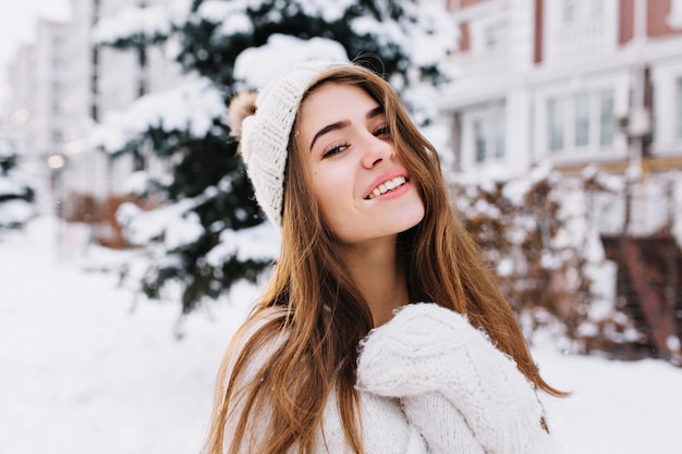 長いブルネットの髪を持つ魅力的な若い女性のスタイリッシュな冬の肖像画。ウールの白い手袋と帽子が雪でいっぱいの通りを歩いています。陽気な気分、笑顔。
