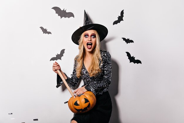 Стильная ведьма в большой шляпе держит тыкву и кричит. Очаровательная блондинка-вампир позирует с осиновым колом.