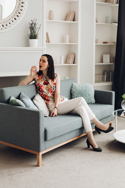 Stylish woman biting marshmallow on couch. casual clothing and heels sitting on modern couch with cushions and biting delicious marshmallow.