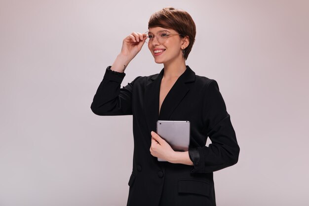 Stylish woman in eyeglasses smiling and holding tablet. Charming lady in black suit poses in good mood on isolated background