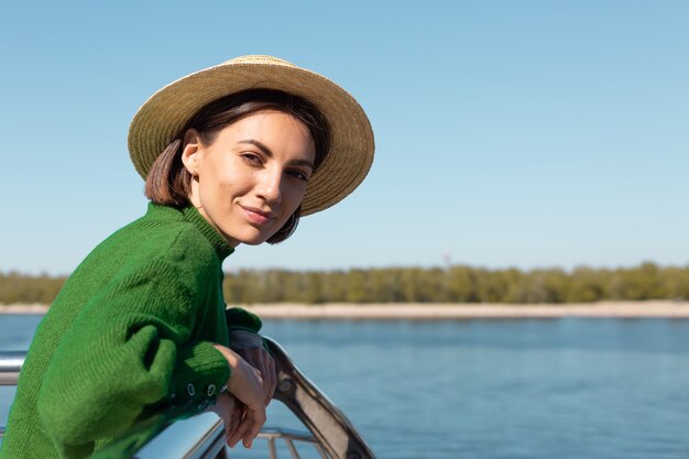 緑のカジュアルなセーターと帽子のスタイリッシュな女性は、川の景色を望む橋の屋外で夏の晴れた日を楽しんでいます