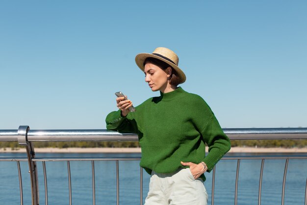 Stylish woman in green casual sweater and hat outdoor on bridge with river view at warm sunny summer day hold mobile phone smile