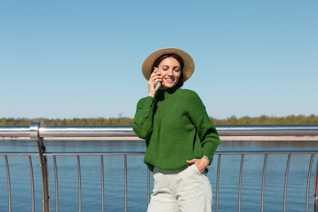 Stylish woman in green casual sweater and hat outdoor on bridge with river view at warm sunny summer day talks on mobile phone