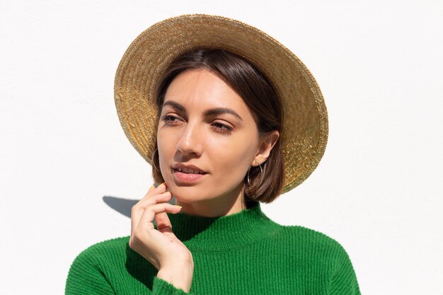 Стильная женщина в зеленом повседневном свитере и шляпе на улице на белой стене со спокойной и уверенной улыбкой