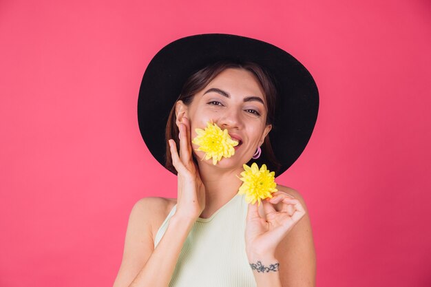 Стильная женщина в шляпе, улыбаясь с двумя желтыми астрами, мило держит один цветок во рту весеннее настроение, счастливые эмоции изолированное пространство