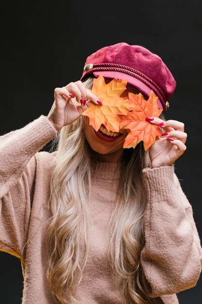 Stylish woman hiding her face behind dry maple leaves against black background