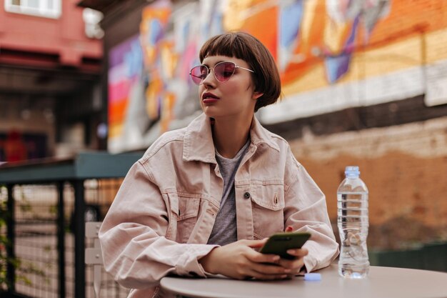 Stylish woman holding smartphone at street Short haired teen in bright glasses and denim beige jacket posing in cafe with bottle water