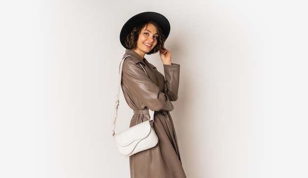 Стильная женщина в кожаном пальто и черной шляпе демонстрирует тенденции зимней моды на белом.