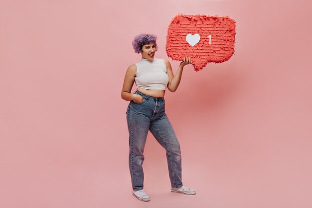 Stylish woman in light clothes and white sneakers posing with red sign like. Bright woman with short hair