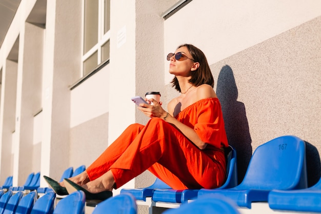 Foto gratuita donna alla moda in abiti arancioni al tramonto allo stadio della pista ciclabile con una tazza di caffè e un telefono cellulare