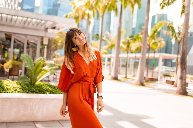 Стильная женщина в оранжевом платье позирует на набережной с пальмами и небоскребами в большом современном городе