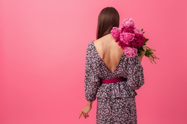 牡丹の花の花束でポーズ夏のトレンディなドレスのピンクの背景にスタイリッシュな女性、後ろからの眺め、セクシーな服