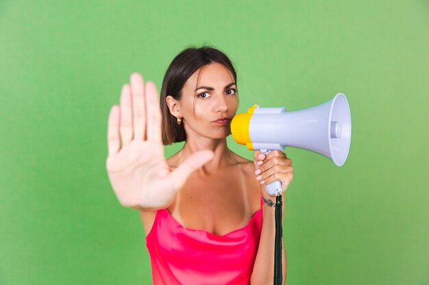 Stylish woman in pink silk dress on green, with megaphone, isolated, show stop gesture with serious face
