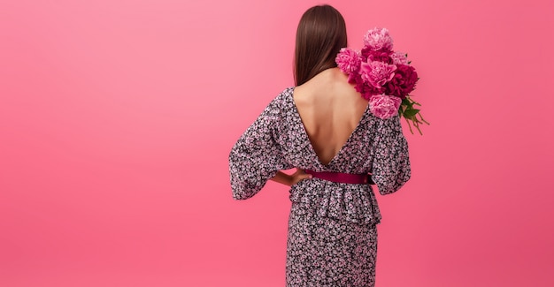 Стильная женщина на розовом в летнем модном платье позирует с букетом цветов пиона, вид со спины