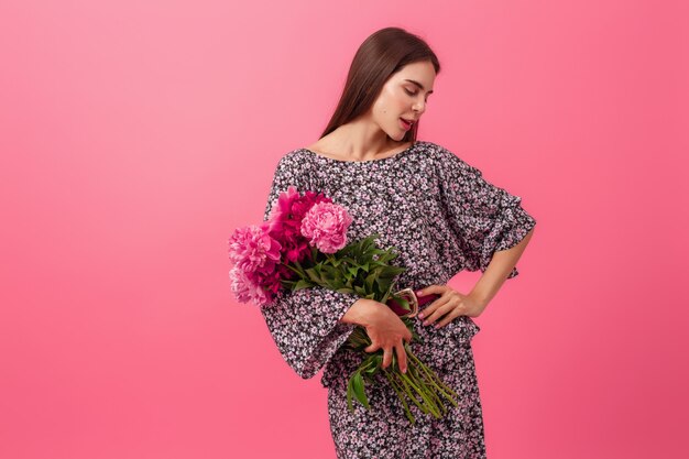 Stylish woman on pink in summer trendy dress posing with peony flowers bouquet