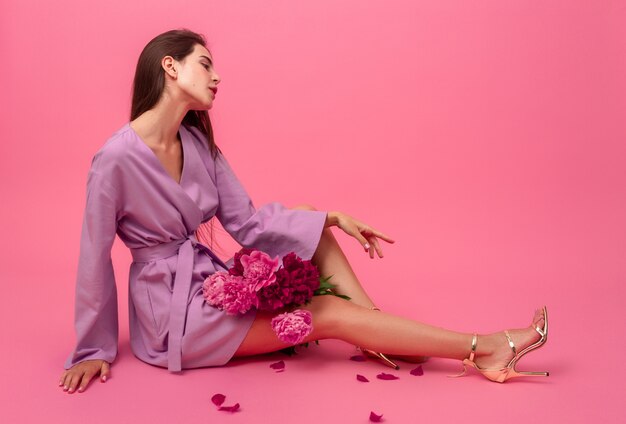 Stylish woman on pink in summer violet mini trendy dress posing with peony flowers bouquet sitting on floor