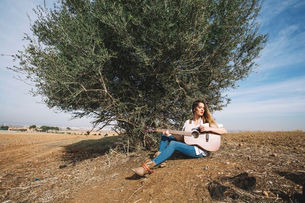 Stylish woman playing guitar near bush