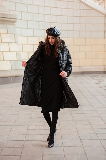 冬の秋のファッショントレンドでポーズをとるスタイリッシュな女性ハイヒールの靴を履いて古い美しい通りで黒のフグコートと革の帽子のベレー帽