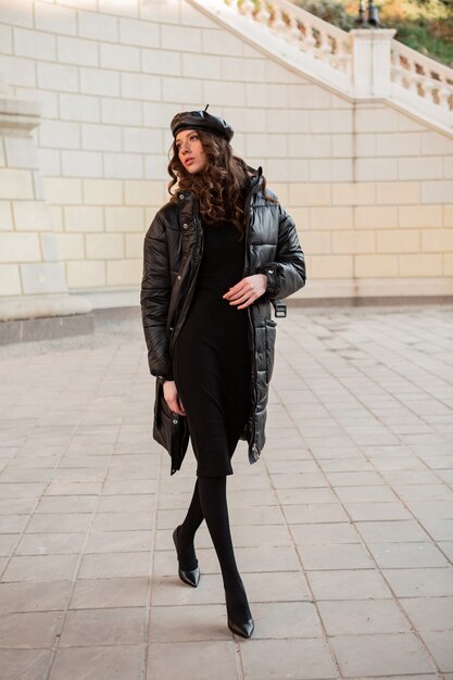 Stylish woman posing in winter autumn fashion trend black puffer coat and leather hat beret in old beautiful street wearing high heel shoes