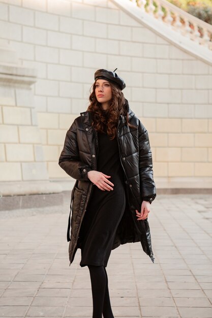 Stylish woman posing in winter autumn fashion trend black puffer coat and leather hat beret in old beautiful street wearing high heel shoes
