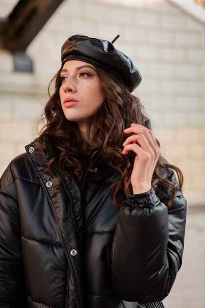 Стильная женщина позирует в зимней осенней модной тенденции черного пуховика и кожаной шляпы в берете на старой красивой улице в туфлях на высоком каблуке