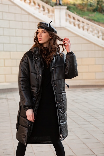 Stylish woman posing in winter autumn fashion trend black puffer coat and leather hat beret in old beautiful street