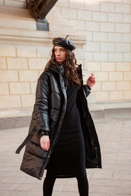 Stylish woman posing in winter autumn fashion trend black puffer coat and leather hat beret in old beautiful street