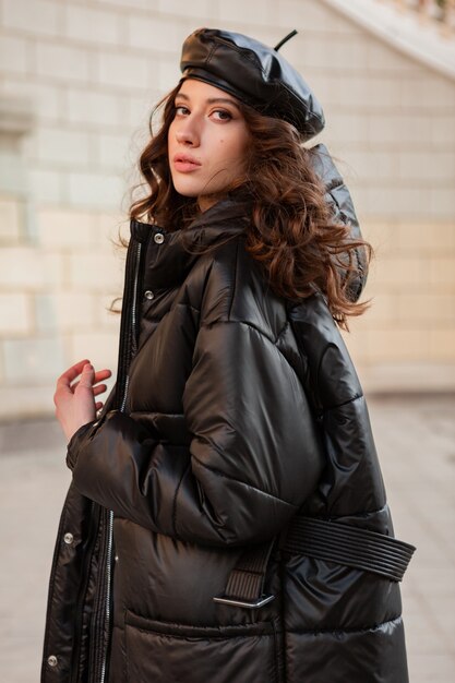 stylish woman posing in winter autumn fashion trend black puffer coat and leather hat beret in old beautiful street