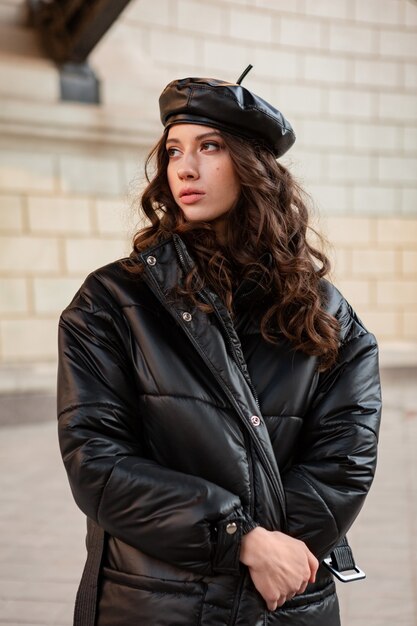 Stylish woman posing in winter autumn fashion trend black puffer coat and leather hat beret in old beautiful street