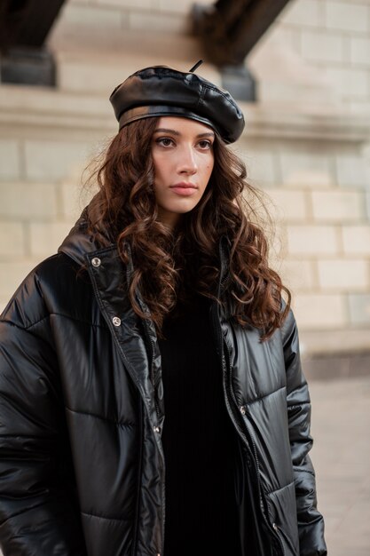 stylish woman posing in winter autumn fashion trend black puffer coat and leather hat beret in old beautiful street