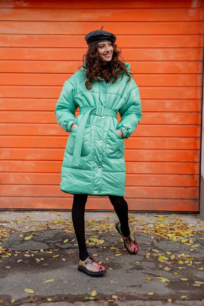 Stylish woman posing in winter autumn fashion trend puffer coat and hat beret against orange wall in street wearing colorful printed shoes