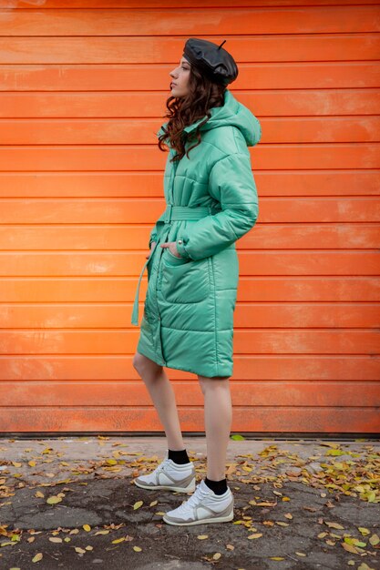 Stylish woman posing in winter autumn fashion trend puffer coat and hat beret against orange wall in street wearing sneakers
