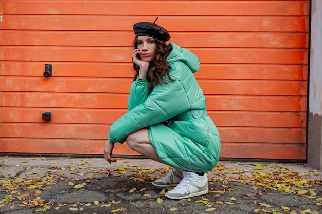Стильная женщина позирует в зимнем осеннем модном пуховике и шляпе-берете на оранжевой стене на улице в кроссовках