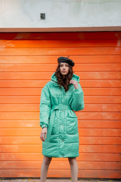Stylish woman posing in winter autumn fashion trend puffer coat and hat beret against orange wall in street