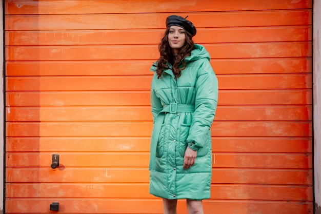 Stylish woman posing in winter autumn fashion trend puffer coat and hat beret against orange wall in street